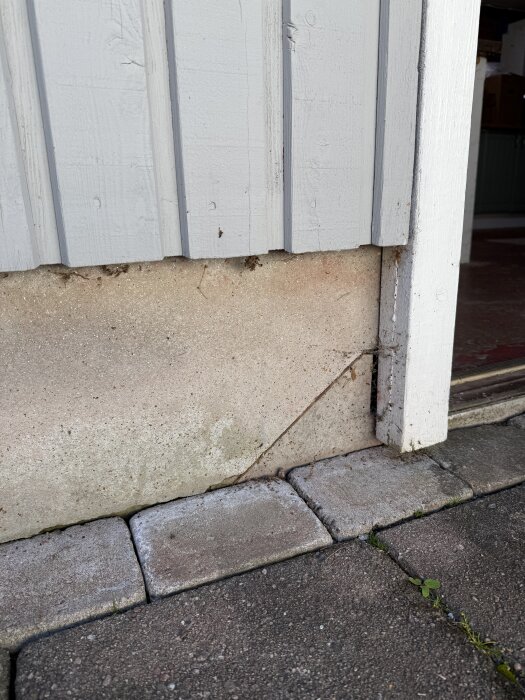 Vägg av träpanel ovanpå en grund av stenskivor med skador, omgiven av markplattor. Skivorna upplevs vara komposit eller stenliknande material.