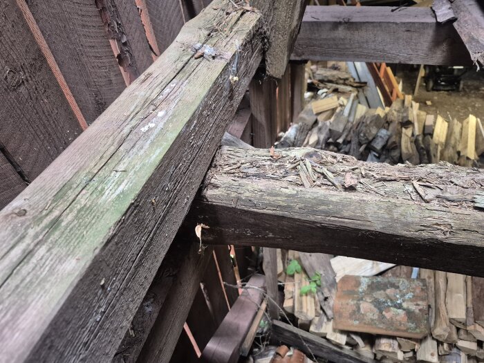 Närbild på slitna och delvis skadade takstolar av trä, omgivna av gamla plankor och bråte i en verkstadsmiljö.