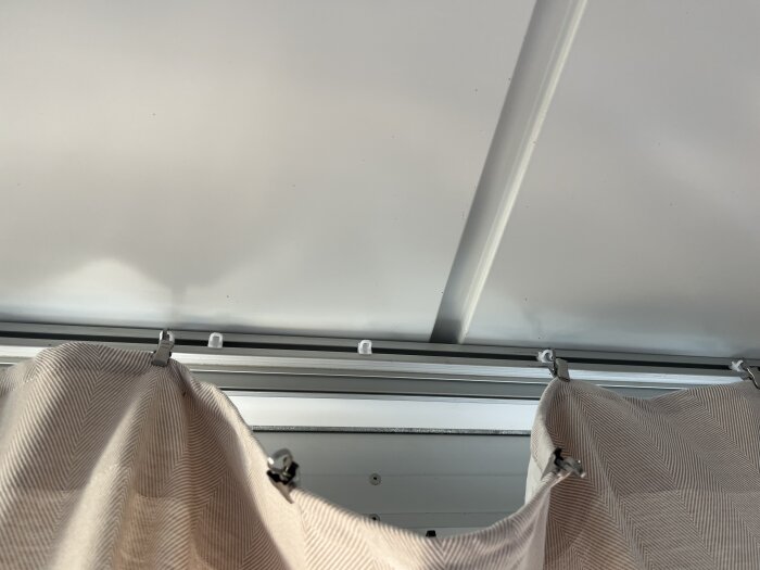 Gardinskena i taket med gardin fäst i skenan, fotograferad från underifrån. Gardinen är upphängd i klämmor som gör att den glider dåligt.
