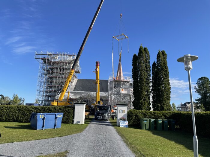 Byggarbetsplats med två kranar som lyfter en tornspira bredvid en byggnadsställning, omgiven av träd och gröna fält under en klarblå himmel.