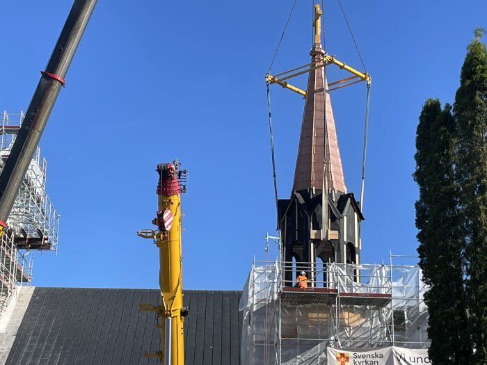 Kyrktorn lyfts med kran under renovering, omgiven av byggnadsställningar och arbetare. Klarblå himmel i bakgrunden.