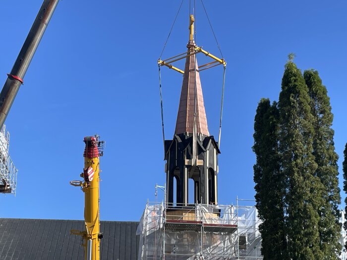 Kyrkospira lyfts med kranar under renovering, blå himmel och träd i bakgrunden.