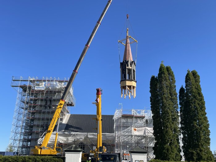 En kran lyfter en kyrktornsspira framför en kyrka med byggnadsställningar, en klar blå himmel i bakgrunden.