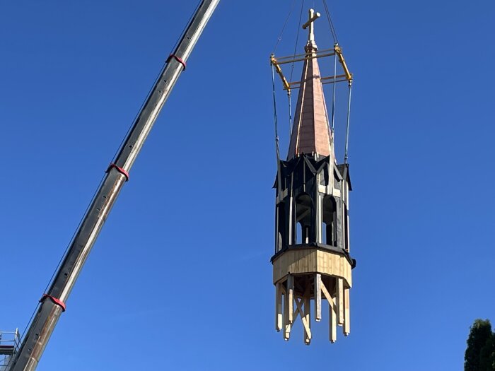 Klocktorn i trä och metall med kopparspira lyfts upp av en kran under klarblå himmel.