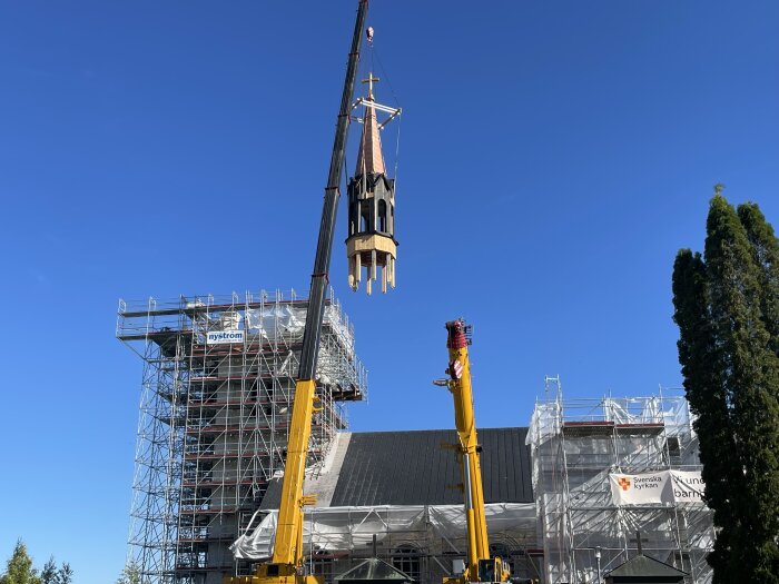 Kran lyfter kyrktorn under renovering medan byggnaden är täckt av byggnadsställningar och presenningar, klarblå himmel i bakgrunden.