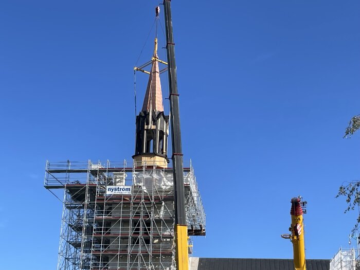 Kyrktorn under renovering, täckt av byggställningar och lyft av en kran mot en klarblå himmel.