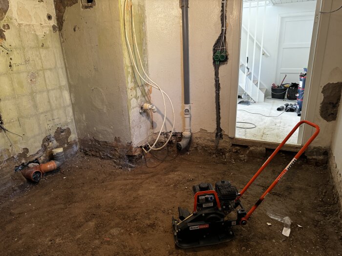 Renovering av badrum visar avlägsnad betong och exponerad rördragning med jordig yta, en komprimeringsplatta på golvet och öppning mot intilliggande rum.