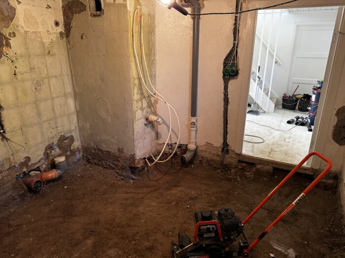 Rivningsarbete i ett badrum med synliga rör och stampad jord, eldosa på väggen. Borttagen betong visar grund för renovering.