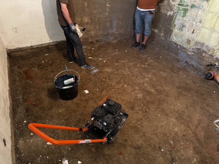 Rum under renovering med borttagen betongplatta, två personer i jeans, en platta, en hink med rulle och en jordvägg synlig.