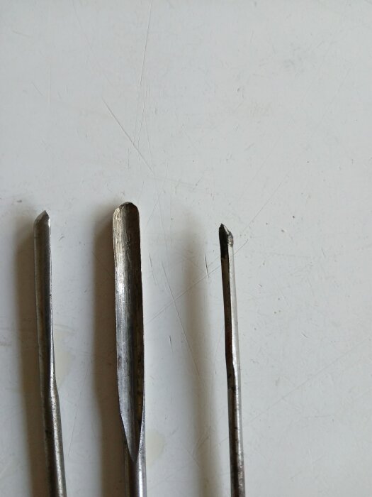 Tre metallverktyg som liknar borrar med varierande spetsig och öppen framände, placerade på en ljus bakgrund.