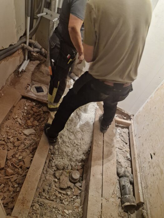 Två personer arbetar i ett badrum med ojämnt underlag, lättbetong appliceras på golvet bredvid rördragningar och träreglar.