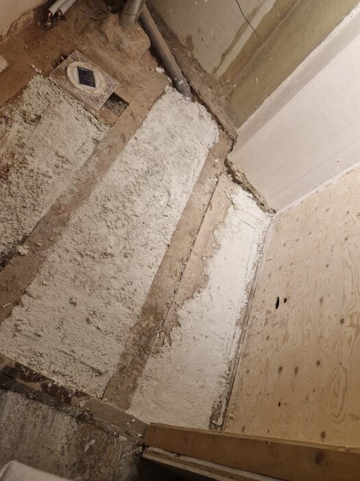 Bilden visar ett badrum under renovering med vit lättbetong applicerad på ett ojämnt golv och mot träpanel, avloppsrör syns också.