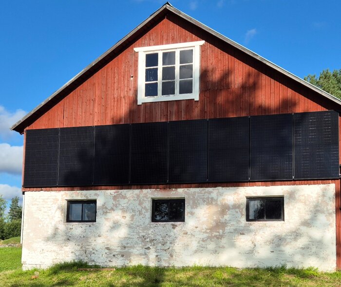 Röd ladugård med solpaneler på övervåningen och vitrinad kalkputsvägg med fönster på nedervåningen. Vackert väder med blå himmel.