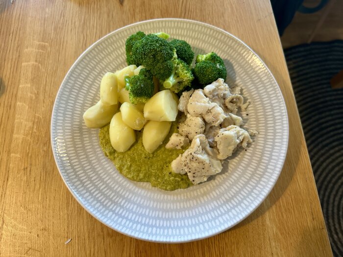 Tallrik med smörstekt kolja, broccoli, potatis och grön ärtsås, serverad på ett träbord.