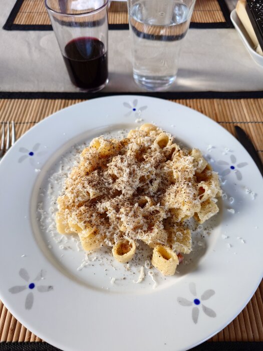 Pasta carbonara serverad på en tallrik med riven ost och peppar, ett glas rött vin och ett glas vatten bredvid.