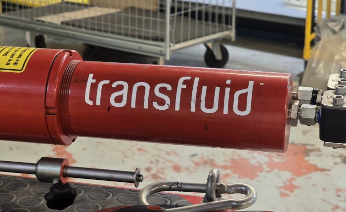 Röd maskindel med texten "transfluid" tryckt på sidan, en del av en industriell verkstadsmiljö med verktyg och utrustning i bakgrunden.