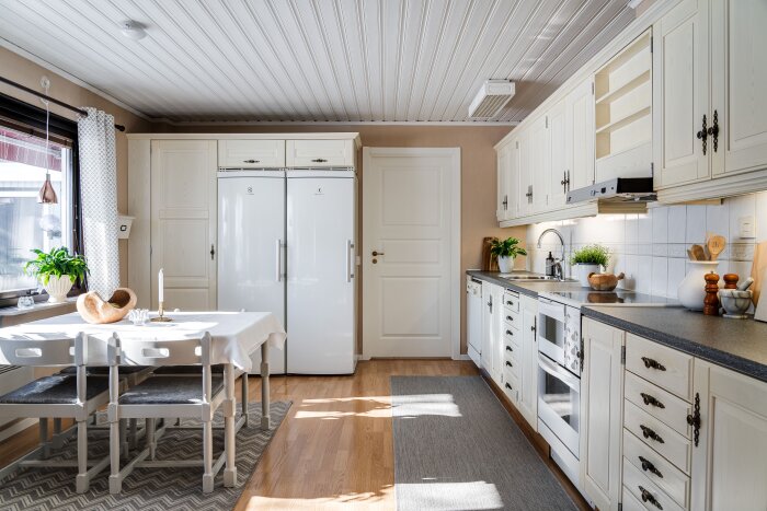 Ljust kök med vita skåpluckor, dubbla kylskåp och matbord. Fönster med gardiner och växter. Ljusinsläpp från vänster.