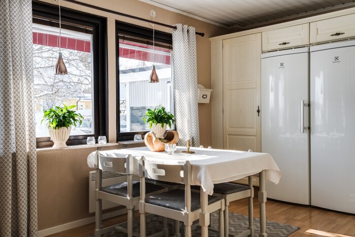 Matplats med vit dukad matbord, fyra stolar, bröd, levande växter på fönsterbräda och kylskåp i bakgrunden. Gardiner med mönster hänger vid fönstren.