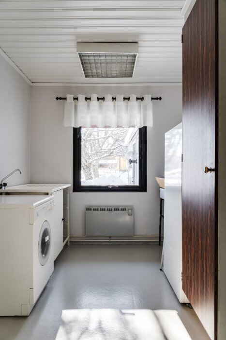Tvättstuga med ett fönster som vetter mot en snöig trädgård, tvättmaskin till vänster, vit vägg och tak med inbyggd belysning.