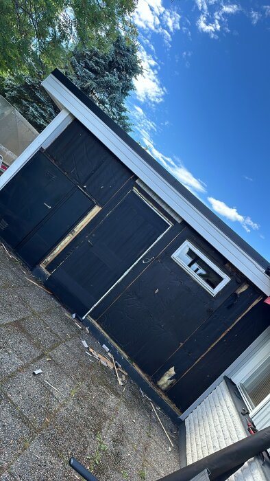 Svartmålad garagevägg med dörr och litet fönster, omgiven av grönska och blå himmel, med bråte på marken framför.
