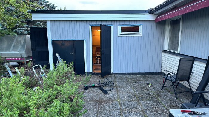 Renoverad del av ett 70-talshus med nymålad panel, öppen dörr, verktyg och grönska på trädgårdsplattor.