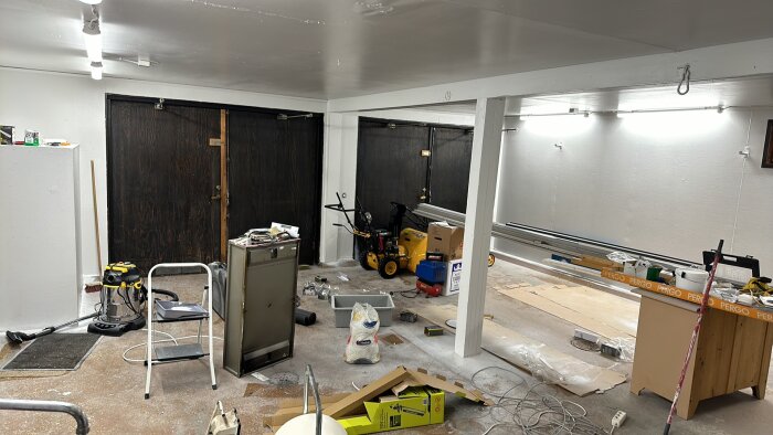 Ett garage under renovering med verktyg, dammsugare, kartonger och byggmaterial utspridda på betonggolvet. Ljusa väggar och mörka dörrar syns.
