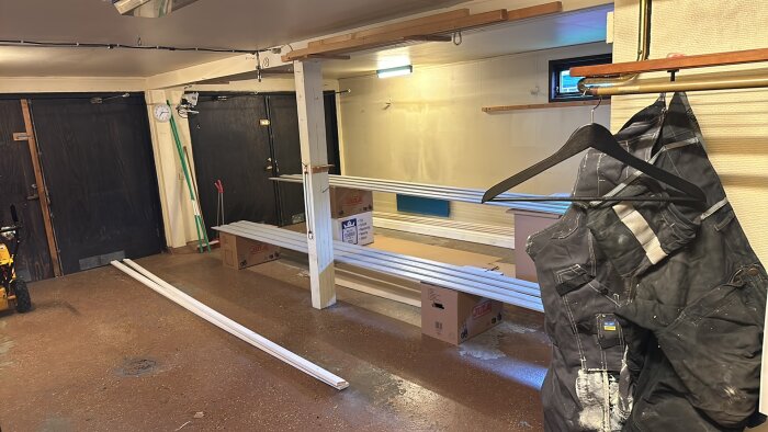 Garage med byggmaterial, kartonger och byggställning. Arbetsoverall hänger bredvid. Vitmålade väggar under renovering.
