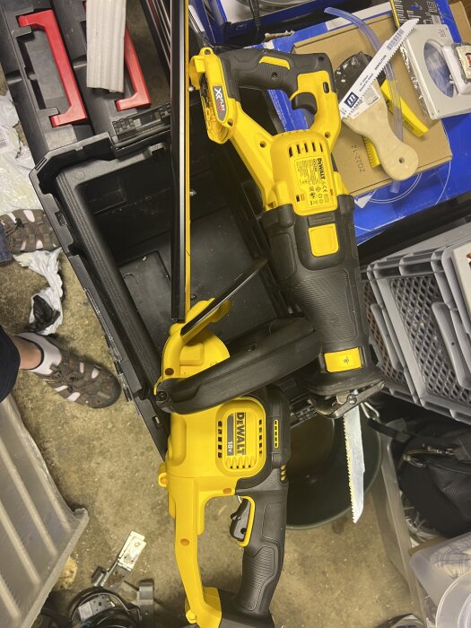 En gul och svart tigersåg med ett sågblad visas i ett stökigt garage, placerad bland verktyg och förpackningar.