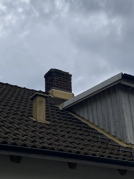 Sliten tegelbeklädd skorsten på tak mot molnig himmel, med synlig takrännor och träpanel av huset intill.