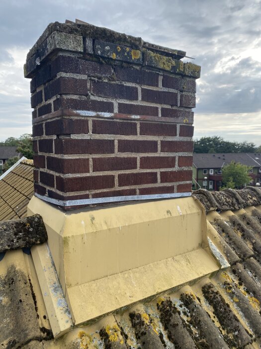 Sliten murad skorsten med sprickor och mossa, placerad på ett tak med gul plåt och tegelpannor med lavar.