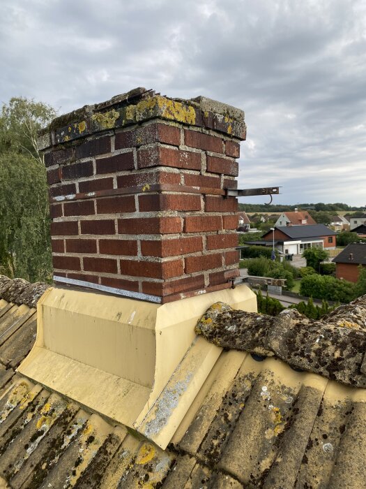 Sliten tegelmurad skorsten med mossa och skadad fogmassa på ett tak, med vy över träd och hus i bakgrunden under molnig himmel.