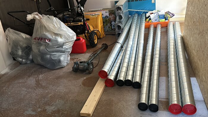 Byggmaterial i garage: ventilationsrör, påsar med delar, en gräsklippare, byggmaterial och en planka på betonggolv.