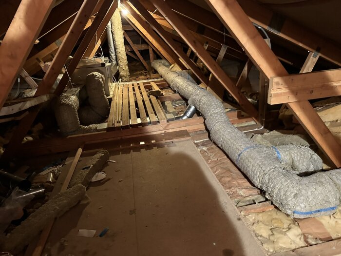Vy av en vind med isoleringsmaterial, ventilationsrör och träreglar som visar pågående renovering och installation av FTX-system.