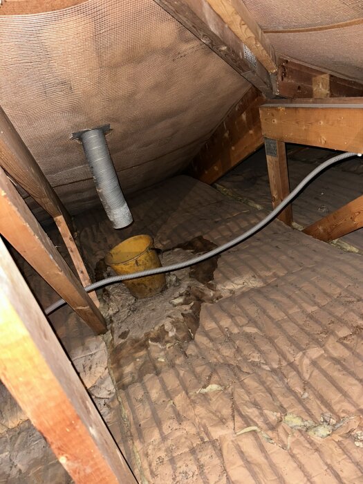 Vind med exponerad isolering, en ventilationsrör och en gul hink placerad på marken, omgiven av träbjälkar.