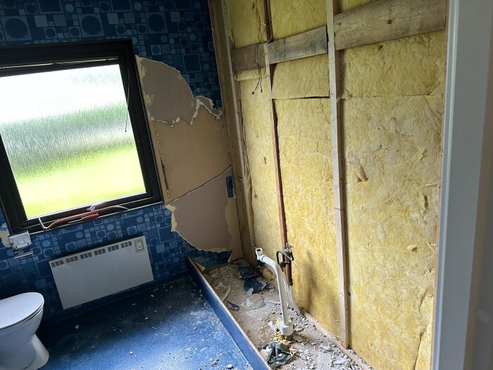 Badrum under renovering med blått golv, halvriven vägg med synlig isolering, fönster bredvid samt toalettstol i vänstra hörnet.