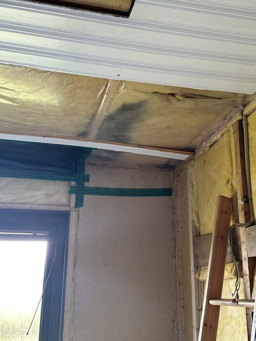 Renovering av ett rum med tillfälligt tejpad plast i taket och blottad isolering, träregel syns i hörnet, nära ett fönster.