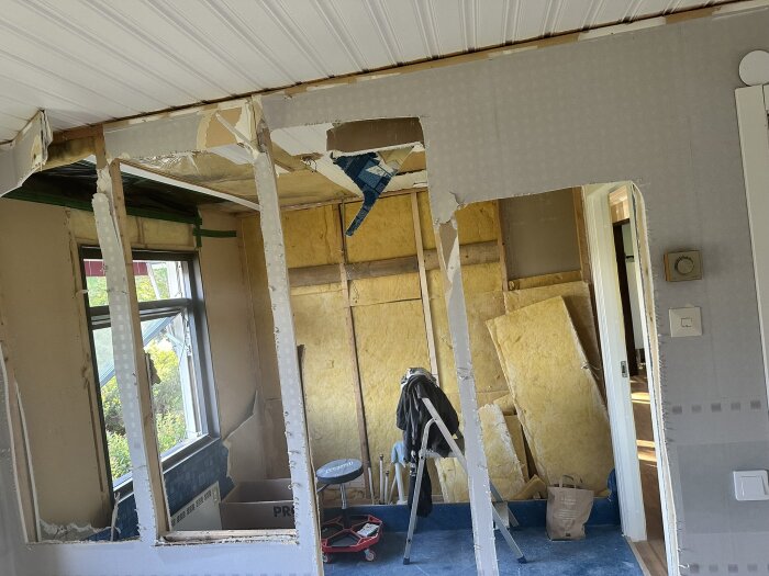 Rivna innerväggar i rum, synlig isolering och verktyg, pågående renovering med byggmaterial och stege.