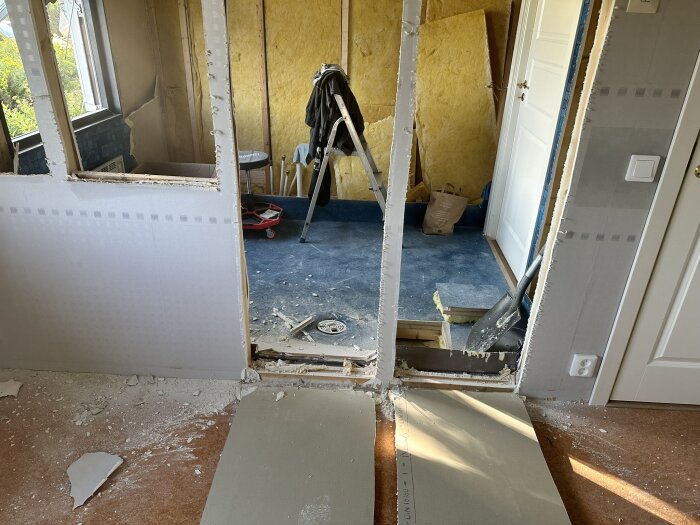 Under renovering, rivna innerväggar och exponerad isolering i ett husprojekt. Verktyg och byggdamm syns på golvet.