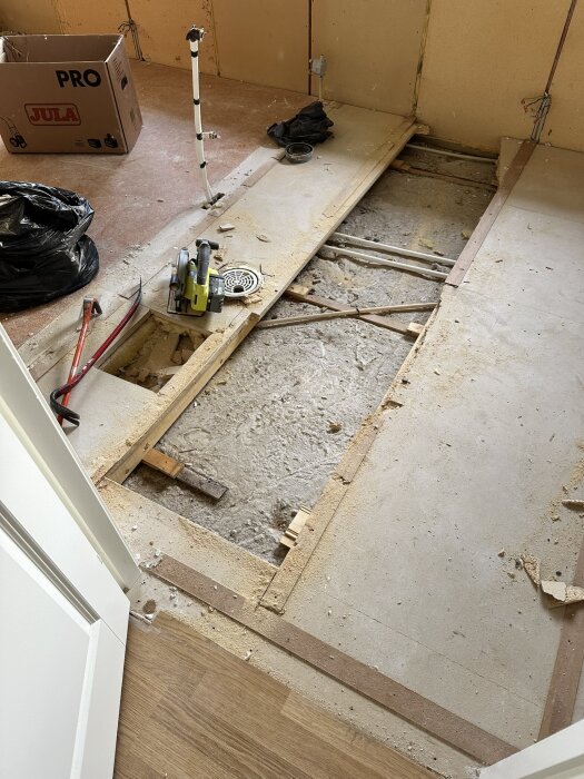Renovering i sovrum där golvet är delvis uppbrutet, verktyg och byggmaterial syns, inklusive en kapsåg och nedbrutet dammigt golv.
