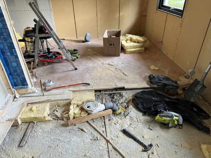 Renoveringsprojekt i ett rum under rivning med verktyg, stege, isolering och öppet golv som avslöjar strukturen under. Kartonger och damm på golvet.