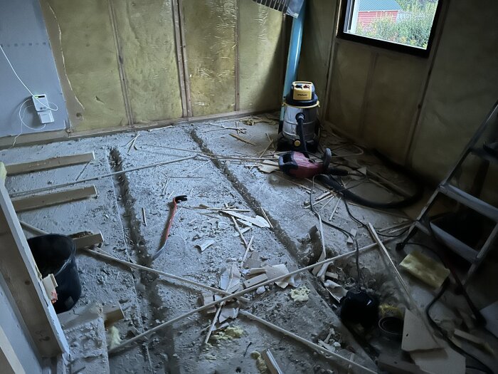 Renovering pågår i ett rum med cementgolv, isolerade väggar och byggmaterial överallt. En dammsugare och verktyg syns på golvet.