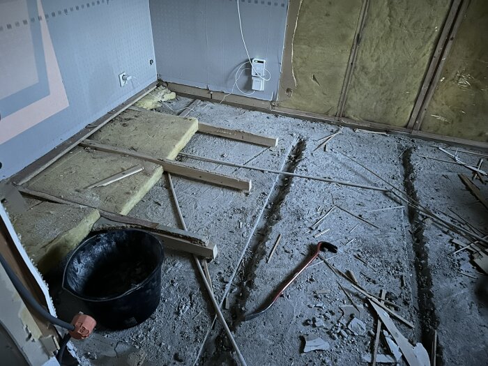 Rivningsarbete i ett rum, golvet är delvis uppbrutet, reglar och isolering synliga, en svart hink och ett verktyg ligger på det dammiga golvet.