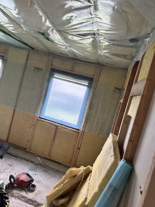 Renoveringsprojekt med blottlagda reglar och isolering i ett rum, plast hänger från taket och en dammsugare ligger på golvet.