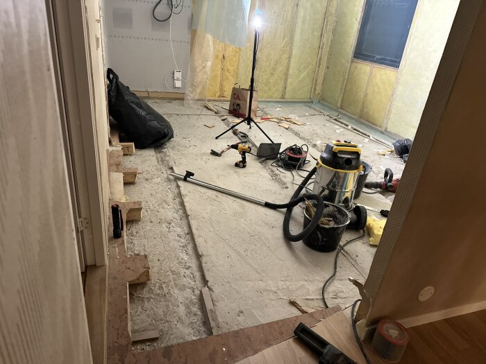 Renoveringsprojekt med utrivet golv och väggar, byggmaterial spritt över rummet, en dammsugare, lampa och verktyg syns i bilden.