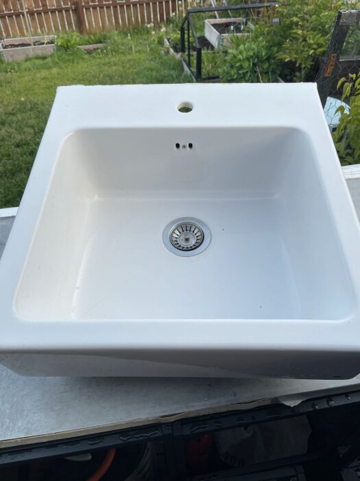 Vit diskho med avrinningshål placerad utomhus på ett bord framför en trädgård.