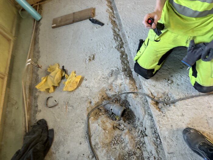 Person i arbetskläder knäböjer vid bilat golv med avloppsrör, omgiven av verktyg och skräp, under badrumsrenovering.