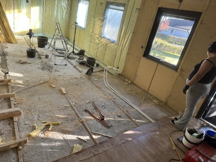 Renovering av rum med öppnade väggar och golv, byggmaterial och verktyg utspridda. Person i förgrunden tittar mot arbetsområdet.