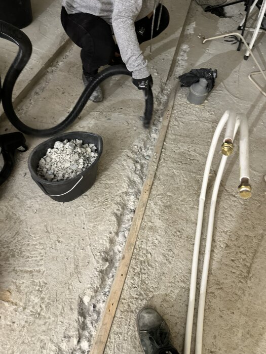 Person använder dammsugare och hink vid renovering, suger upp betongrester från ett uppbilat avloppsrör i betonggolvet. Golvplatta och rör syns.