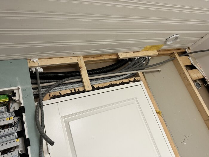 Elektrisk central med automatsäkringar och slangar i tak och vägg under renovering, delvis dold av dörrkarm.
