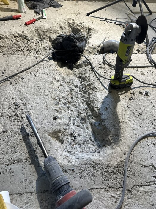 Översiktsbild av pågående golvbilning för avloppsrör, med verktyg som vinkelslip och borr i förgrunden. Rubble och kablar syns.